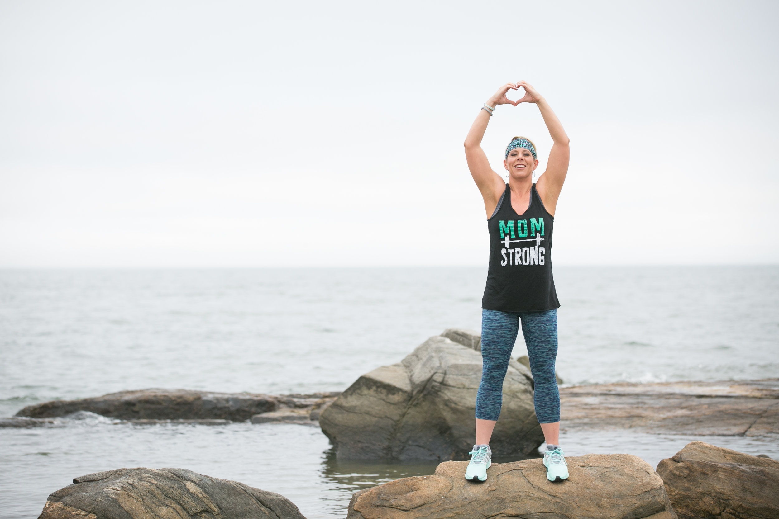 fit mom on rocks at beach with hands in heart&nbsp;